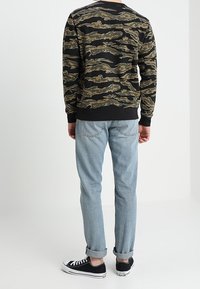 Svart sweatshirt med ett camouflagemönster i nyanser av grönt och brunt, tillsammans med ljusblå jeans med uppvikta ärmar och svarta sneakers.