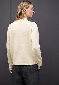 Pull en tricot couleur crème avec un col haut et un ourlet côtelé ; texture douce, manches longues, vu de dos sur fond sombre.