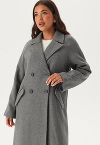 Cappotto doppiopetto in misto lana grigio con rever a notched, grandi tasche frontali e chiusura a bottoni. Texture morbida, vestibilità fluida.