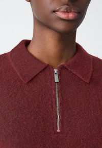 Pull-over en mélange de laine bordeaux avec un col à revers et une fermeture éclair à l'avant. Le tissu présente une texture douce avec un motif subtil.
