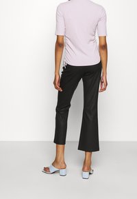 Persona in piedi contro uno sfondo bianco semplice, che indossa un top arricciato viola chiaro, pantaloni neri a vita alta e sandali con tacco blu chiaro.