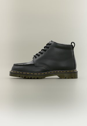 Schwarzer Leder-Knöchelstiefel mit schwarzen Schnürsenkeln, gelber Naht auf dicker Sohle und einer Zuglasche hinten, seitlich auf beige Hintergrund gezeigt.
