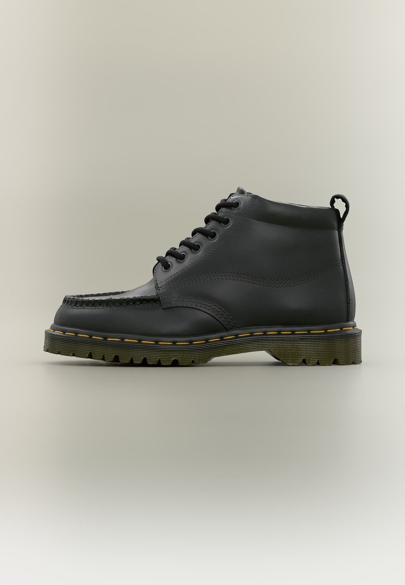 Schwarzer Leder-Knöchelstiefel mit schwarzen Schnürsenkeln, gelber Naht auf dicker Sohle und einer Zuglasche hinten, seitlich auf beige Hintergrund gezeigt.
