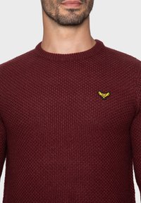 Bordeauxroter Strickpullover mit einer strukturierten, gerippten Optik und rundem Ausschnitt. Verfügt über ein kleines gelbes, gesticktes Logo auf der Brust.