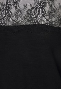 Schwarzes Oberteil mit einem zarten Spitzeneinsatz am Ausschnitt, aufwendigen floralen Mustern und einem gewellten Saum an der Spitze. Weiche Textur.