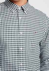 Camisa de cuadros verde y blanca con cuello, que presenta botones blancos redondeados y un pequeño logo en el lado izquierdo del pecho.
