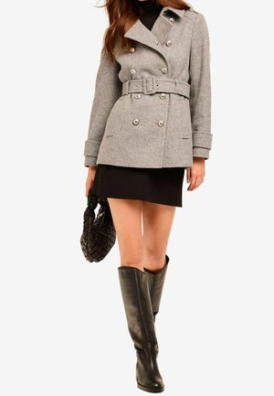 Femme portant un manteau gris ceinturé à double boutonnage, une mini-jupe noire, des bottes noires montant au genou et tenant un sac à main noir tissé.