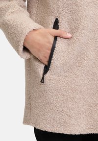 Beige Fleece-Mantel mitstrukturierter Oberfläche, ausgestattet mit einer seitlichen Reißverschlusstasche und einem lässigen Schnitt. Der Kragen ist weich und rund.