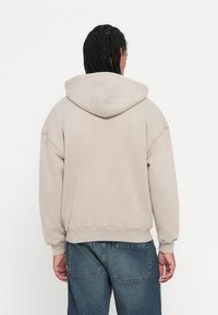 Hoodie bege claro com um capuz grande, punhos e barra em canelado. Tecido suave e texturizado. Usado sobre jeans de denim azul claro.