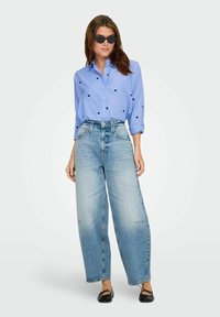 Šviesiai mėlyna dryžuota marškiniai su juodais taškais, derinami su plačiomis šviesiai mėlynomis džinsomis ir juodais plokščiais batais. Aksesuarai – juodi akiniai nuo saulės.