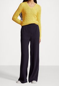 Gul kabelstickad tröja med V-halsdesign, tillsammans med vida marinblå byxor. Detaljer inkluderar en liten logotyp på tröjan.