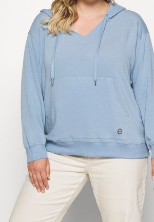 Femme portant un sweat à capuche bleu clair à col en V avec poche avant et un pantalon de couleur crème, debout sur un fond blanc uni.