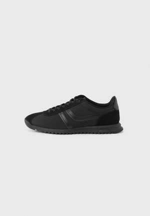 Chaussure de sport noire basse avec lacets à l'avant, deux bandes latérales et semelle texturée sur fond gris clair uni.