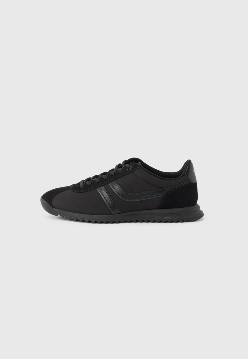 Chaussure de sport noire basse avec lacets à l'avant, deux bandes latérales et semelle texturée sur fond gris clair uni.