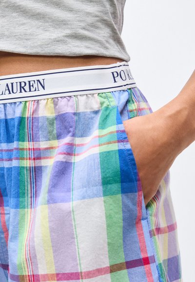 Άτομο που φοράει πολύχρωμα καρό σορτς με το χέρι στην τσέπη και γκρι πουκάμισο εισερχόμενο ελαφρώς πάνω από τη ζώνη με την ετικέτα "Polo Ralph Lauren."