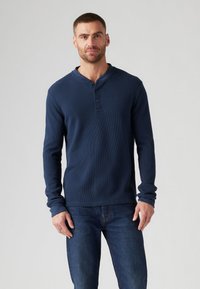 Sötétkék henley póló, bordázott anyagból, három gombos kivágással, hosszú ujjakkal és fitted fazonnal, farmerkabáttal párosítva.