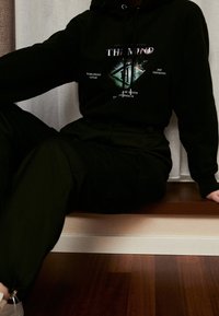 Černá mikina s kapucí s grafickým textem a diamantovým tvarem, spárovaná s tmavými kalhotami. Hladká látka, volný střih, manžety na rukávech s kroužky a elastický pas.