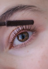 Brun læbe med lange, mørke vipper, et subtilt rosa øjenlåg og et øjenbryn; en mascara-børste børster mod vippen.