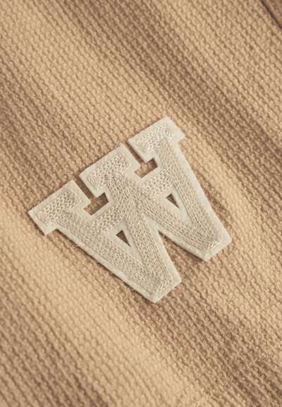Tissu texturé beige avec un logo "W" brodé en blanc en relief. Le matériau présente un motif tissé avec des détails subtils.