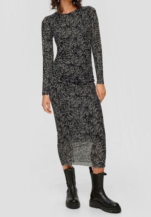 Robe noire à manches longues avec un superposé transparent à motif abstrait blanc, silhouette ajustée et fentes à l'ourlet, assortie à des bottes noires.
