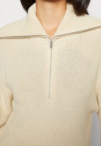 Femme portant un pull crème en maille avec un large col et une demi-fermeture éclair à l'avant, visible du cou à la taille.