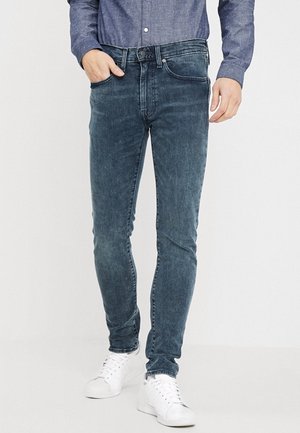 Mann trägt dunkelblaue, eng anliegende Jeans, ein blaues Hemd mit Knöpfen, weiße Sneaker, mit einer Hand in der Tasche vor einfarbigem Hintergrund.