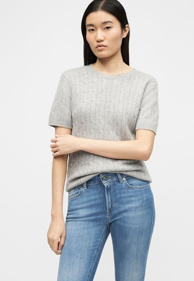 Νεαρή γυναίκα με ίσια σκούρα μαλλιά φορώντας ένα ανοιχτό γκρι κοντομάνικο πουλόβερ με πλεκτό σχέδιο και ανοιχτόχρωμο τζιν παντελόνι, με σταυρωμένα χέρια.