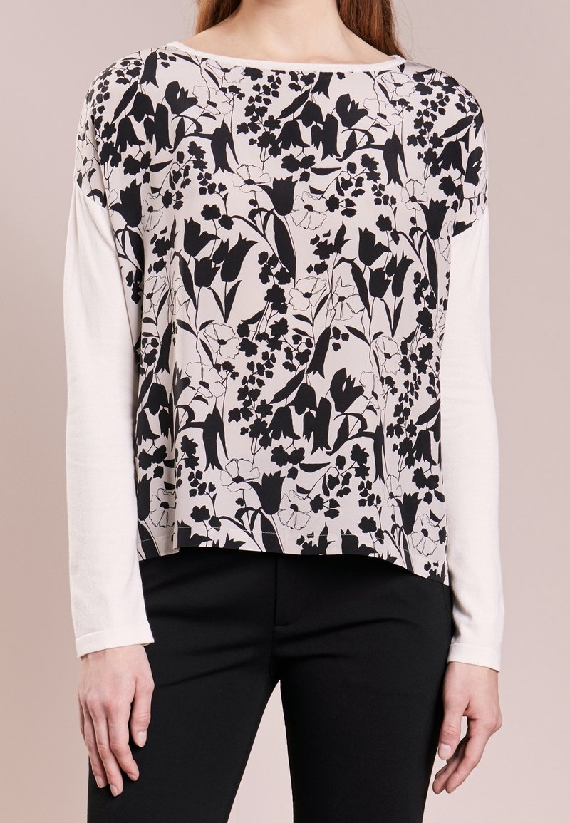 Długi rękaw top z czarnym kwiatowym wzorem na beżowym tle, z gładką białą okrągłą szyją i obrzeżem. Gładka tekstura tkaniny.