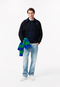 Giacca bomber blu navy su un maglione blu navy, jeans azzurri e scarpe da ginnastica bianche. Tiene una sciarpa a righe blu e verde.
