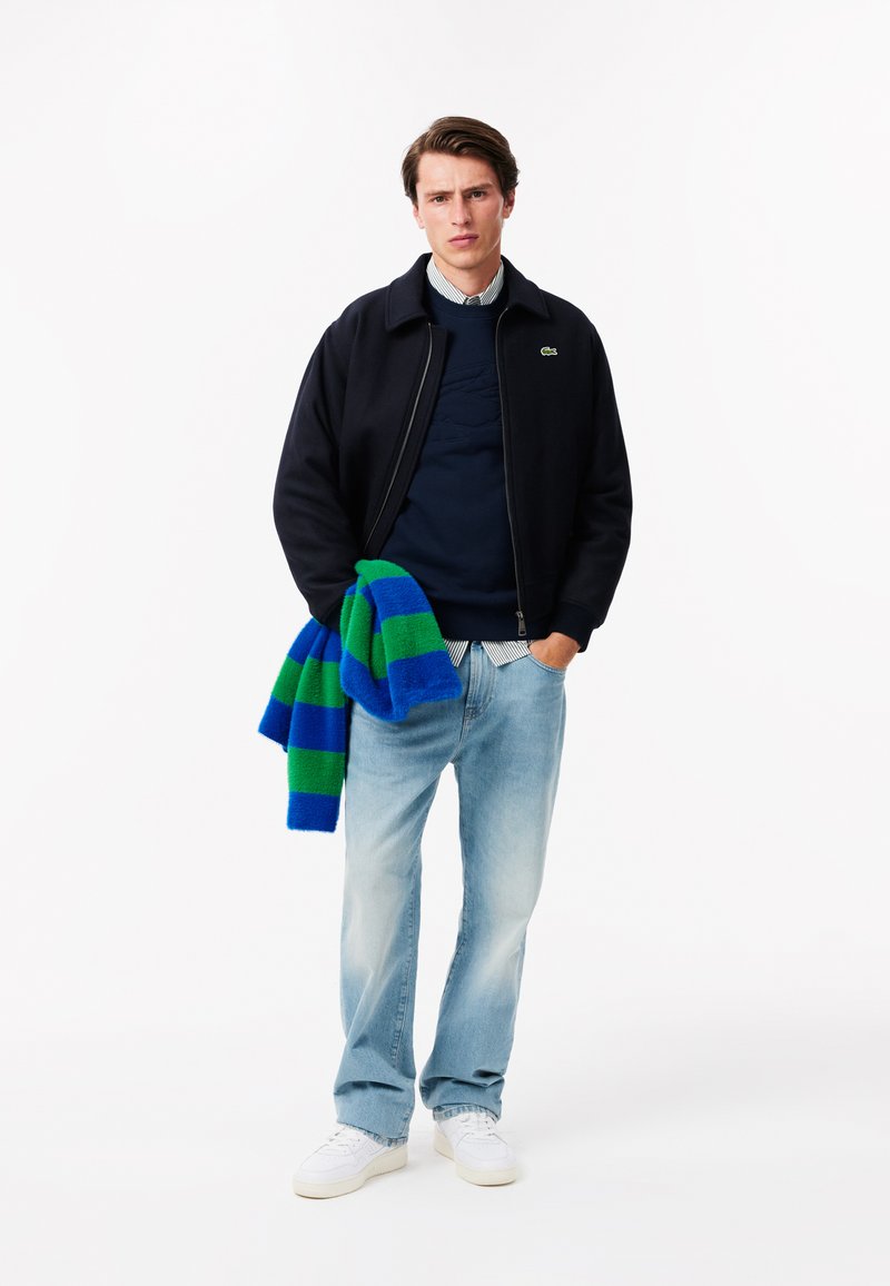 Granatowa kurtka bombowiec na granatowym swetrze, jasnoniebieskie dżinsy i białe trampki. Trzyma niebiesko-zieloną w paski szalik.