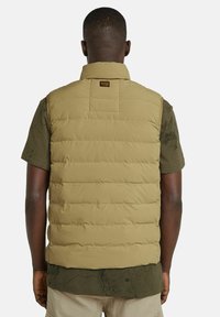 Chaleco acolchado color caqui con costuras horizontales, cuello alto y una etiqueta cuadrada en la parte posterior; textura ligera y suave, diseño sin mangas.