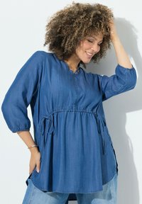 Blusa in denim con scollatura rotonda, maniche a 3/4 e vita elastica. Presenta lacci in vita e fessure laterali. Tessuto blu, texture liscia.