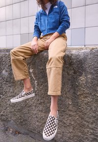 Cappuccio blu tagliato, pantaloni beige a vestibilità ampia e scarpe slip-on a scacchi bianco e nero. Superficie di pietra grigia testurizzata sullo sfondo.