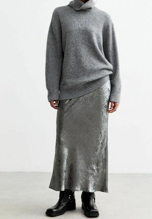 Person iført en grå rullekrave, metallisk sølvfarvet ankel-lang nederdel og sorte ankelstøvler, stående mod en ensfarvet baggrund.