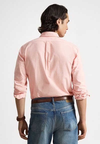 Roza majica z dolgimi rokavi, zavihanimi rokavi, zadnjim delom s šivom in malim naborkom, kombinirana s svetlo modrimi kavbojkami in rjavim pasom.