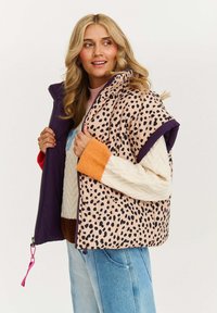 Bezrękawnik puchowy z beżowego materiału w czarne cętki lamparta, fioletowymi wykończeniami i różowym suwakem. Nosi się go na dzianinowy sweter.