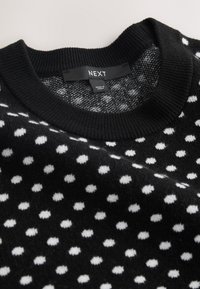 Pull noir en maille avec des pois blancs et un col côtelé, avec une étiquette cousue portant la mention "NEXT" et "Made in China."