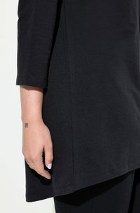 Tunic nera a maniche lunghe con orlo curvo. Tessuto morbido, presenta dettagli di cuciture leggere e una texture liscia. Braccio visibile con tatuaggio.