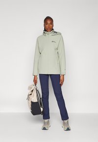 Light green waterproof jacket with a hood, paired with navy pants and gray hiking shoes. Holding a beige and black backpack.