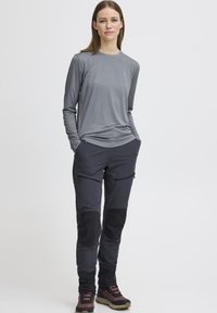 Grijze top met lange mouwen, gecombineerd met zwarte broek met meerdere zakken en kniestukken. Dragen met robuuste, meerkleurige wandelschoenen.