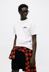 Νέος άνδρας που φοράει μαύρα γυαλιά με πλαίσιο, λευκό μπλουζάκι "Obey Studios", μαύρο σορτς και κόκκινο-μαύρο καρό πουκάμισο δεμένο γύρω από τη μέση του.