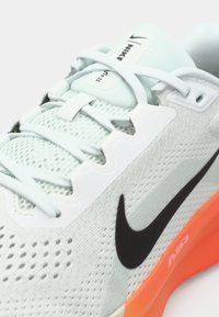 Zapatilla deportiva Nike gris claro con parte superior de malla, logo Swoosh negro, detalles blancos y suela vibrante naranja. Presenta cordones planos y un diseño perforado.