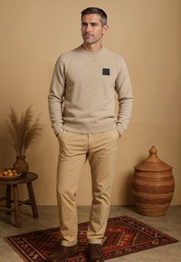 Lichtbeige trui met een zwart patch, gecombineerd met kaki broek. Bruine schoenen. De achtergrond bevat een decoratief tapijt en aardse decoratieve elementen.