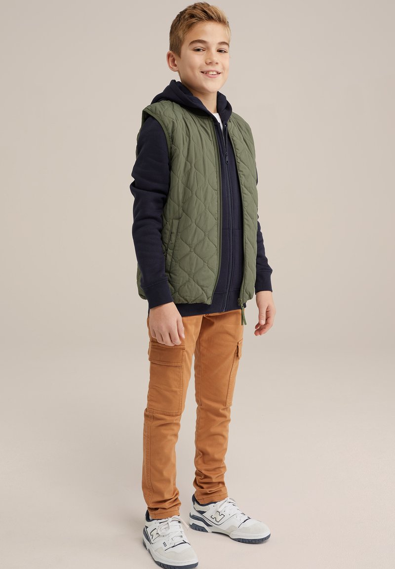 Groene gewatteerde vest over een marineblauwe hoodie, gecombineerd met tan cargobroeken en witte sneakers met marineblauwe accenten. Eenvoudige, casual stijl met zakken.