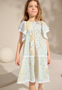 Hellblaues Blumenkleid mit Rüschensäumen, mehrlagigem Design und weißer Spitzenverzierung. Die Stoffe zeigen unterschiedliche Blumen- und Karomuster.