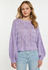 Lila gestrickter Pullover mit lockerer Passform, ausgeschnittenen Mustern, gerippten Bündchen und runder Ausschnitt, kombiniert mit hellblauen Jeans.