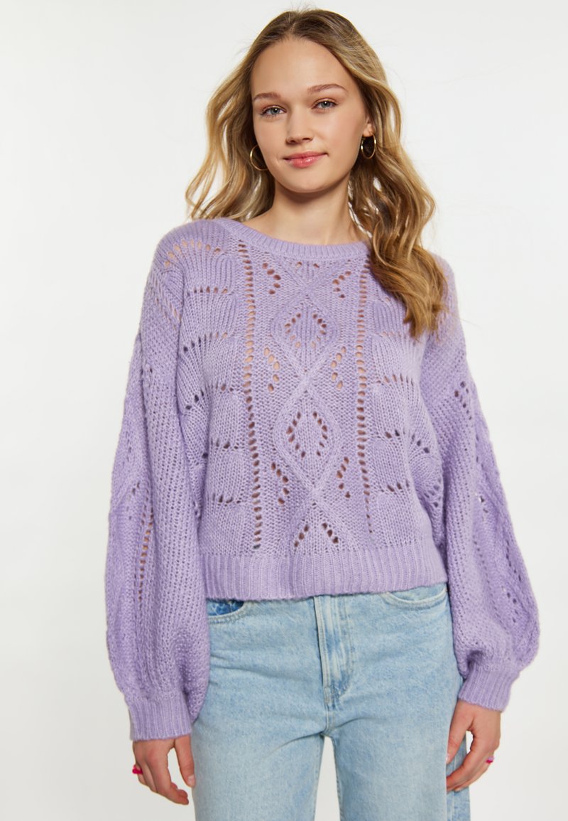 Lila gestrickter Pullover mit lockerer Passform, ausgeschnittenen Mustern, gerippten Bündchen und runder Ausschnitt, kombiniert mit hellblauen Jeans.