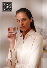 Pot de soin en verre avec un couvercle rose doré, tenu par une personne portant une chemise boutonnée beige clair. Arrière-plan flou aux tons neutres.