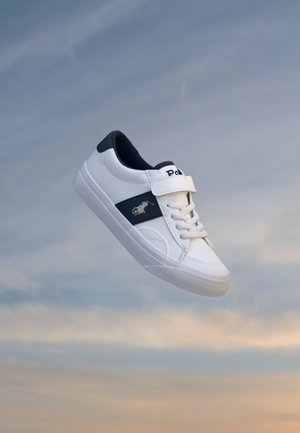 Baskets blanches et bleu marine avec bride en velcro et lacets flottant devant un ciel nuageux.