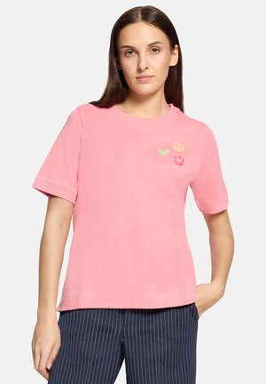 Donna con una maglietta rosa a maniche corte con piccole icone di cuore, simbolo di pace e faccine sorridenti sul petto, abbinata a pantaloni blu navy a righe sottili.